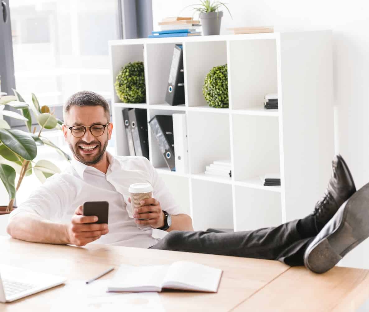 Image of cheerful handsome man in formal wear sitting in office with legs at table, and using black smartphone
